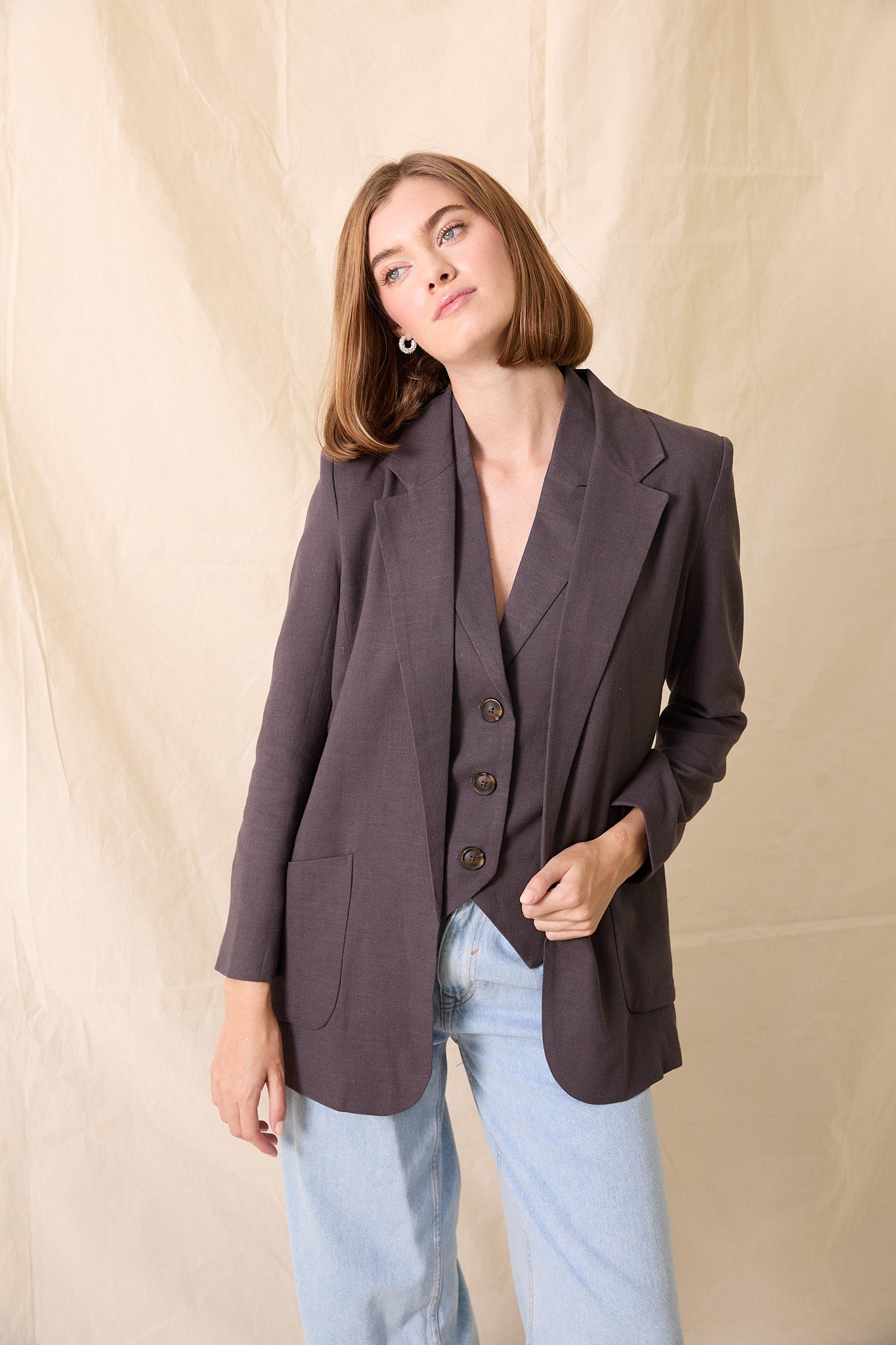 Woman wearing a dark brown blazer over a shirt with buttons, standing against a beige background