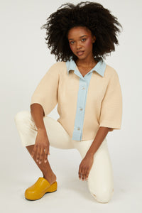 Woman wearing a beige sweater over a light blue shirt with white pants on a white background